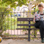 Stephanie, große Brille, blass, sitzt mit einem Laptop auf einer Bank in einem Park, dem Conertplatz. Neben ihr ein blau-gelber Ball.