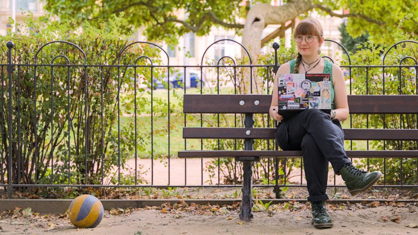 Stephanie, große Brille, blass, sitzt mit einem Laptop auf einer Bank in einem Park, dem Conertplatz. Neben ihr ein blau-gelber Ball.