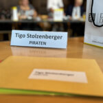 Namensschild mit dem Namen Tigo Stolzenberger darauf auf einem Tisch. Davor grpße Briefumschläge.